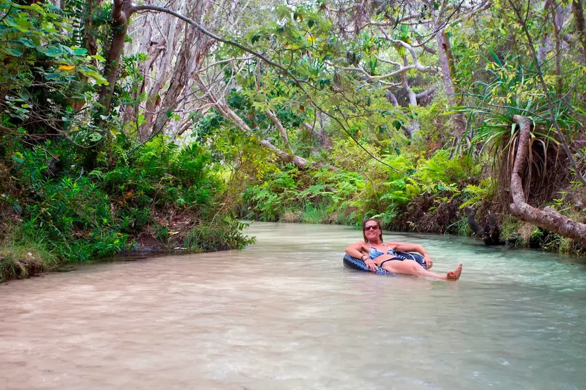 Woman relaxing on an inflatable tube in the clear shallow water of Eli Creek on K’gari Fraser Island, surrounded by lush greenery Title