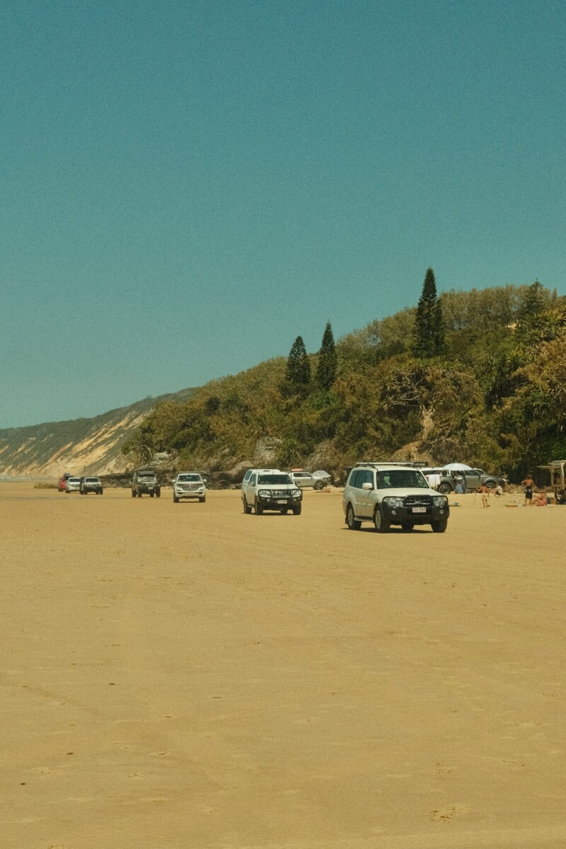 Dundee Adventure Travel | Where To Park In Rainbow Beach Before Your K’gari (Fraser Island) Tour 4WD tag-along tour convoy driving on 75 Mile Beach on K’gari (Fraser Island) with blue sky and ocean backdrop.