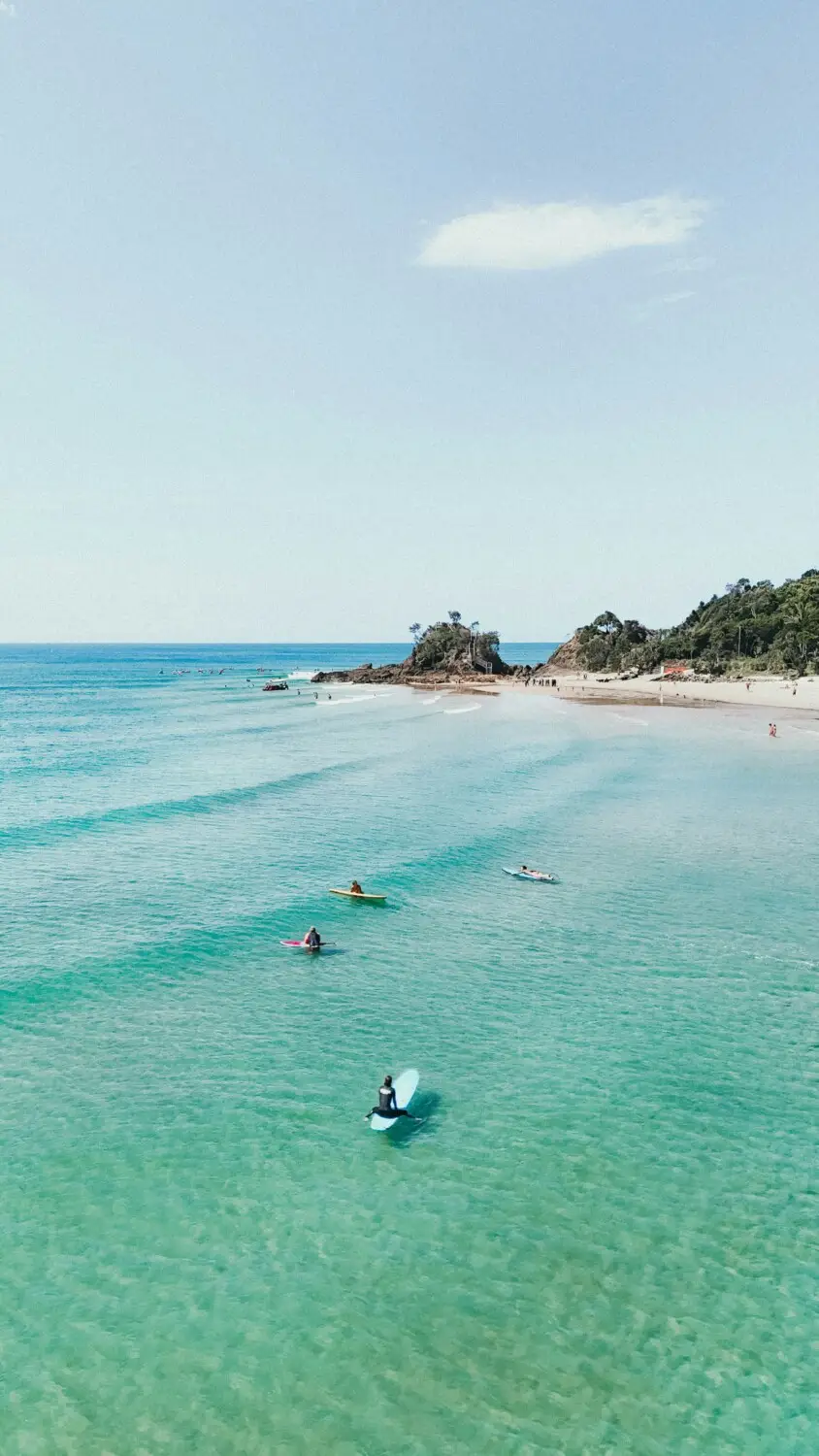 Surfers paddling in clear turquoise water near a sandy beach and rocky headland on a sunny day