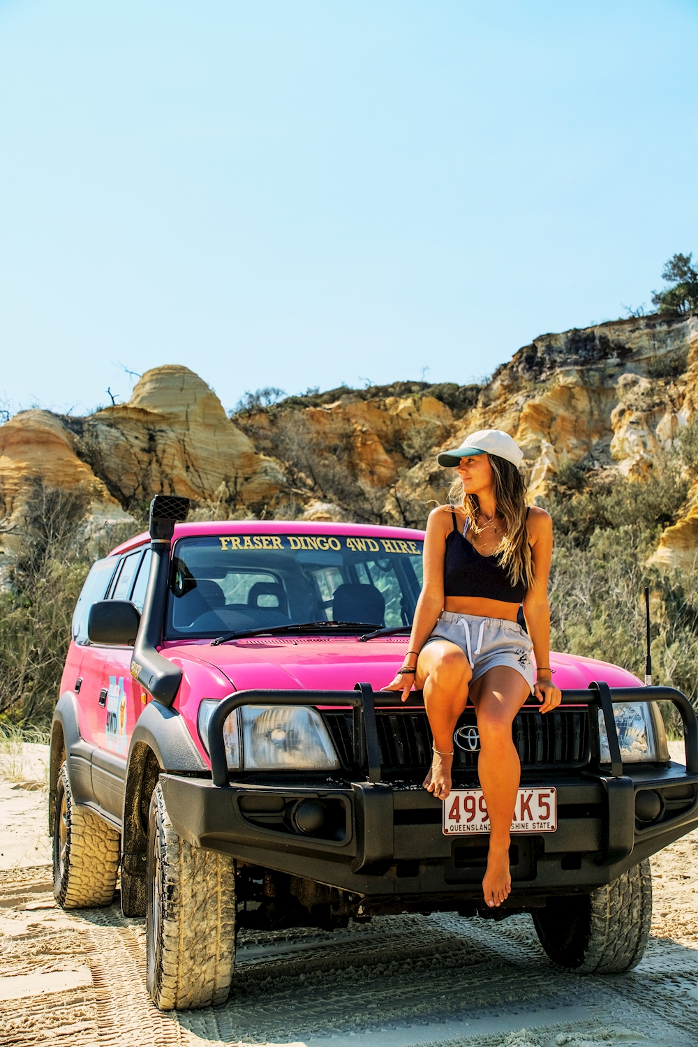 Woman sitting on front of pink 4WD with sand cliffs behind on Fraser Island
